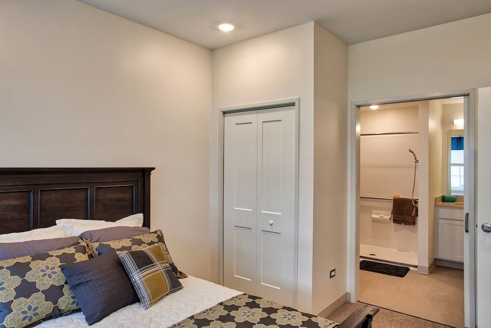 A cozy bedroom with a neatly made bed, accented with patterned pillows and a view of the closet and adjacent room.