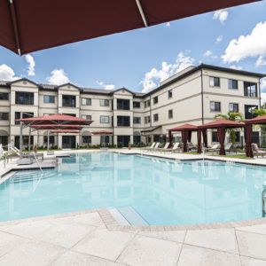 A modern apartment complex featuring a spacious outdoor swimming pool with loungers and shaded areas under a clear blue sky.