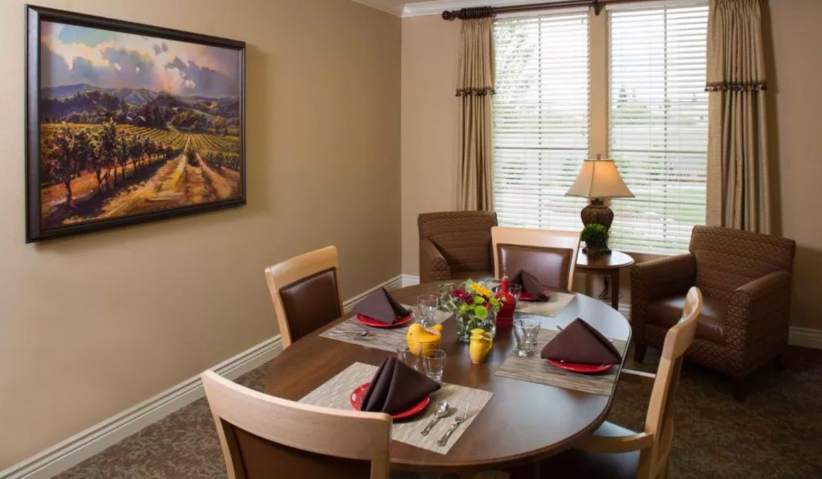 A cozy dining room setup with a round table adorned with napkins and flowers, situated near a window with a lamp and wall artwork.