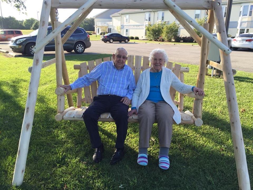 An elderly couple sitting together on a wooden swing in a sunny park.