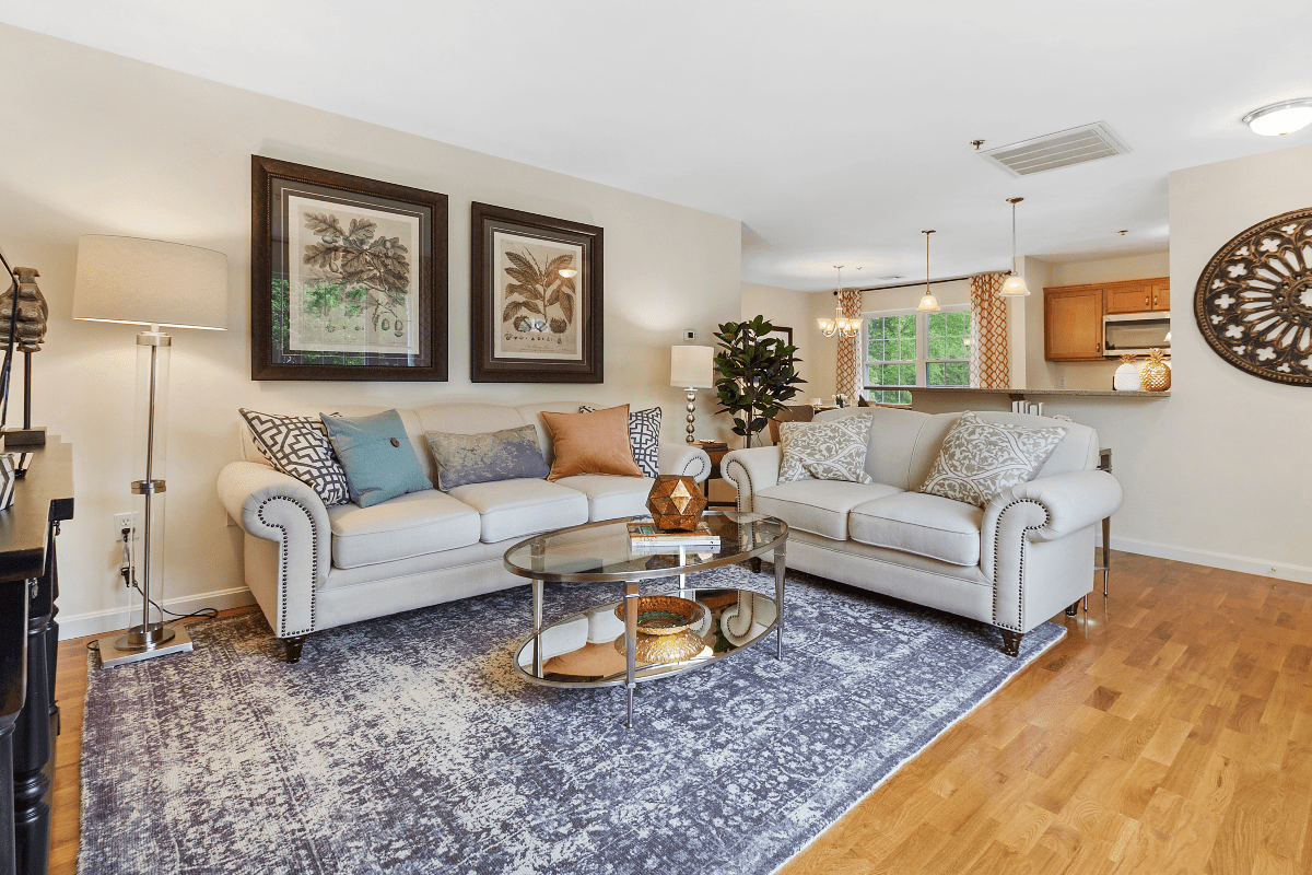 A cozy and elegant living room featuring two sofas, a glass coffee table, decorative pillows, artistic paintings, and ambient lighting.