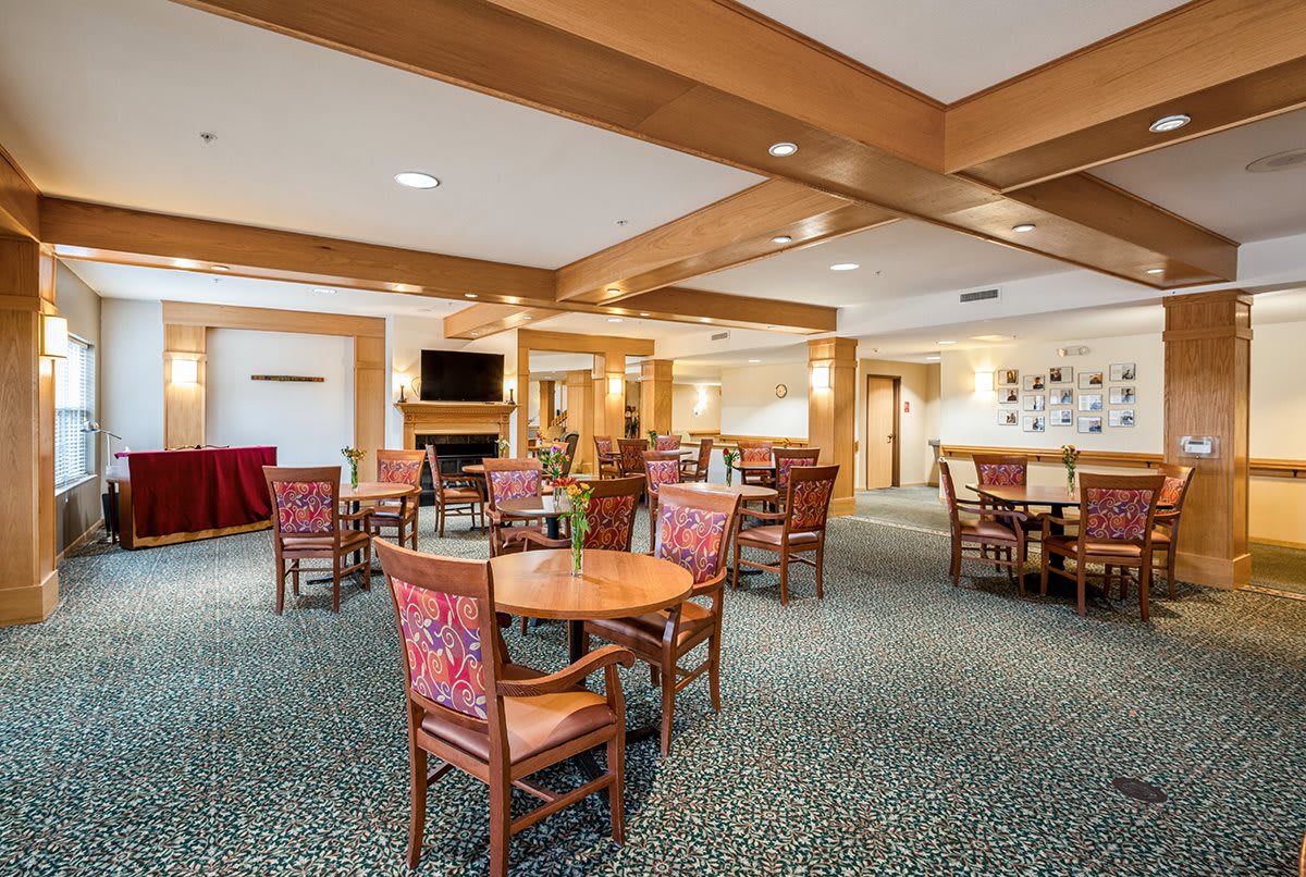 A cozy dining area with wooden beams, tables, and chairs arranged on a carpeted floor.