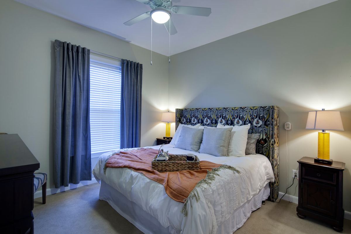 A cozy bedroom with a king-sized bed, decorative pillows, and a soft blanket. The room features a window with blue curtains and a bedside lamp.