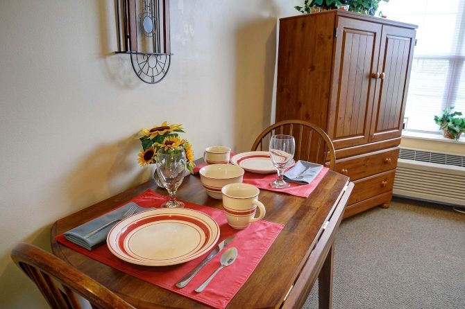 A dining table set for two with plates, cups, and cutlery on red placemats, accompanied by a vase of flowers.