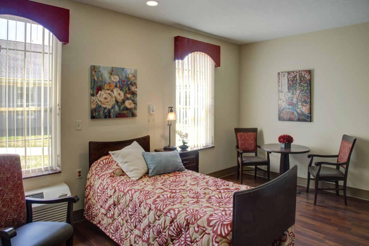A cozy bedroom with a single bed adorned with a patterned red comforter, two pillows, and flanked by artwork on the walls. The room features a small table with chairs and a window allowing natural light.
