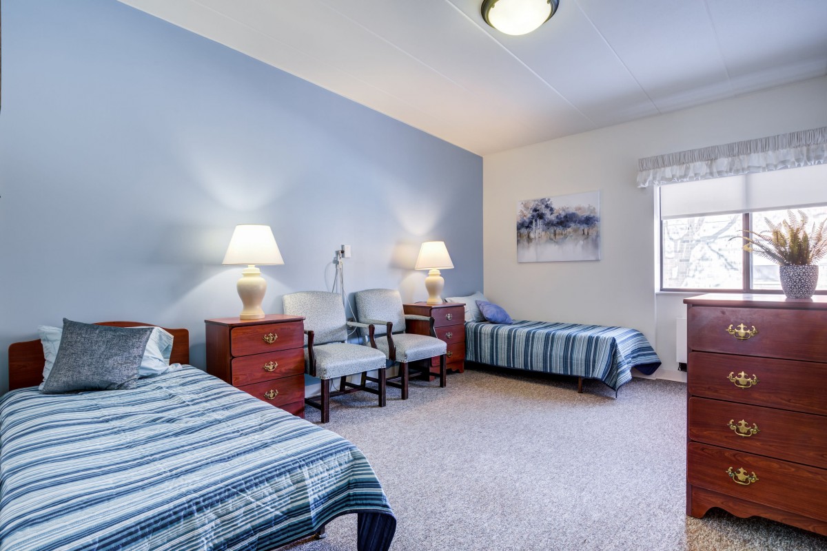 A cozy bedroom with twin beds, blue bedding, and elegant nightstands with lamps, complemented by chairs and wall art.
