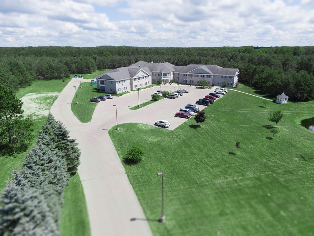 An aerial view of a building surrounded by greenery, featuring a parking lot and a road under a cloudy sky.