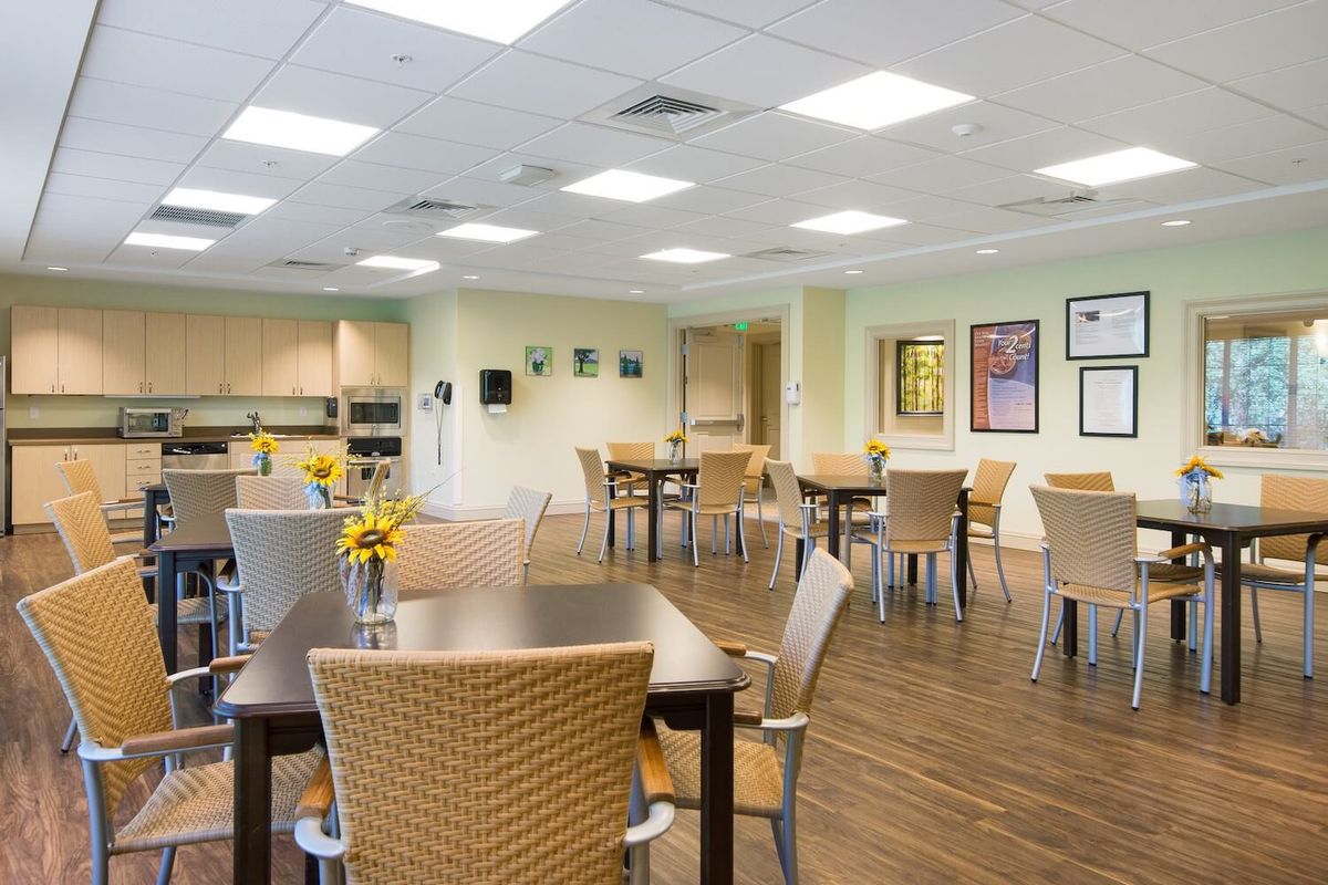 A modern dining area with several tables and wicker chairs, featuring bright lighting and a kitchenette in the background.