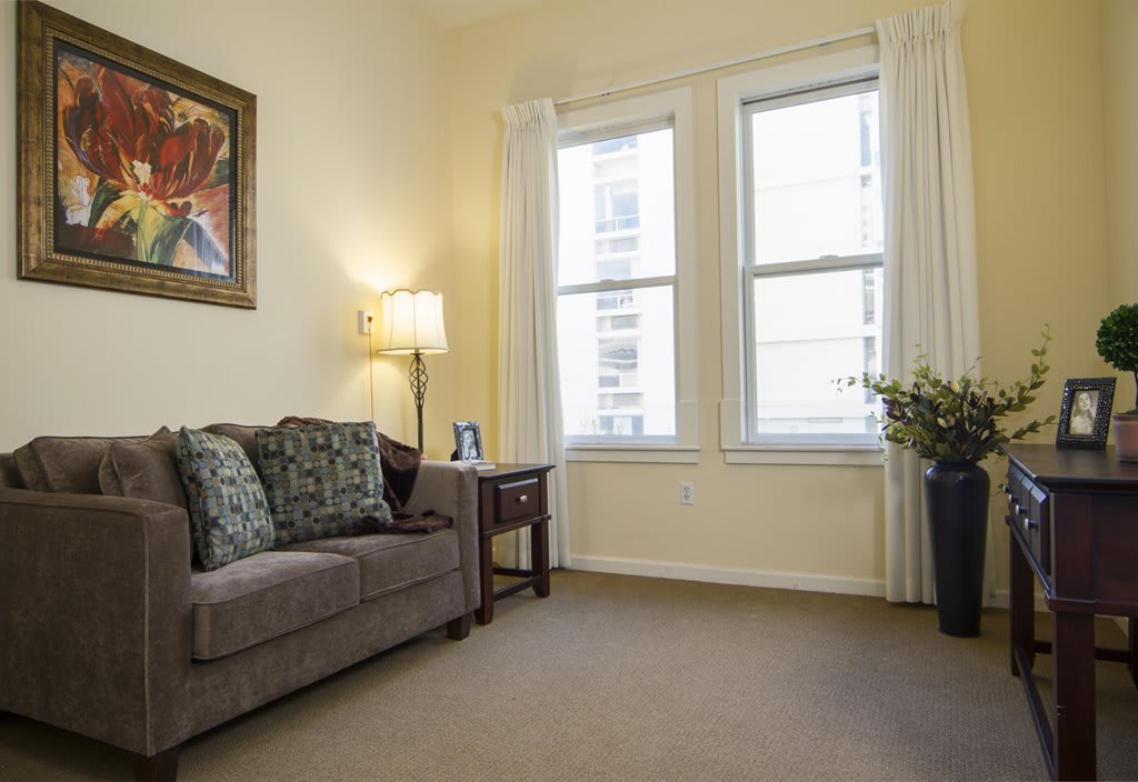 A cozy living room with a comfortable sofa, a side table with a lamp, large windows, and an indoor plant.