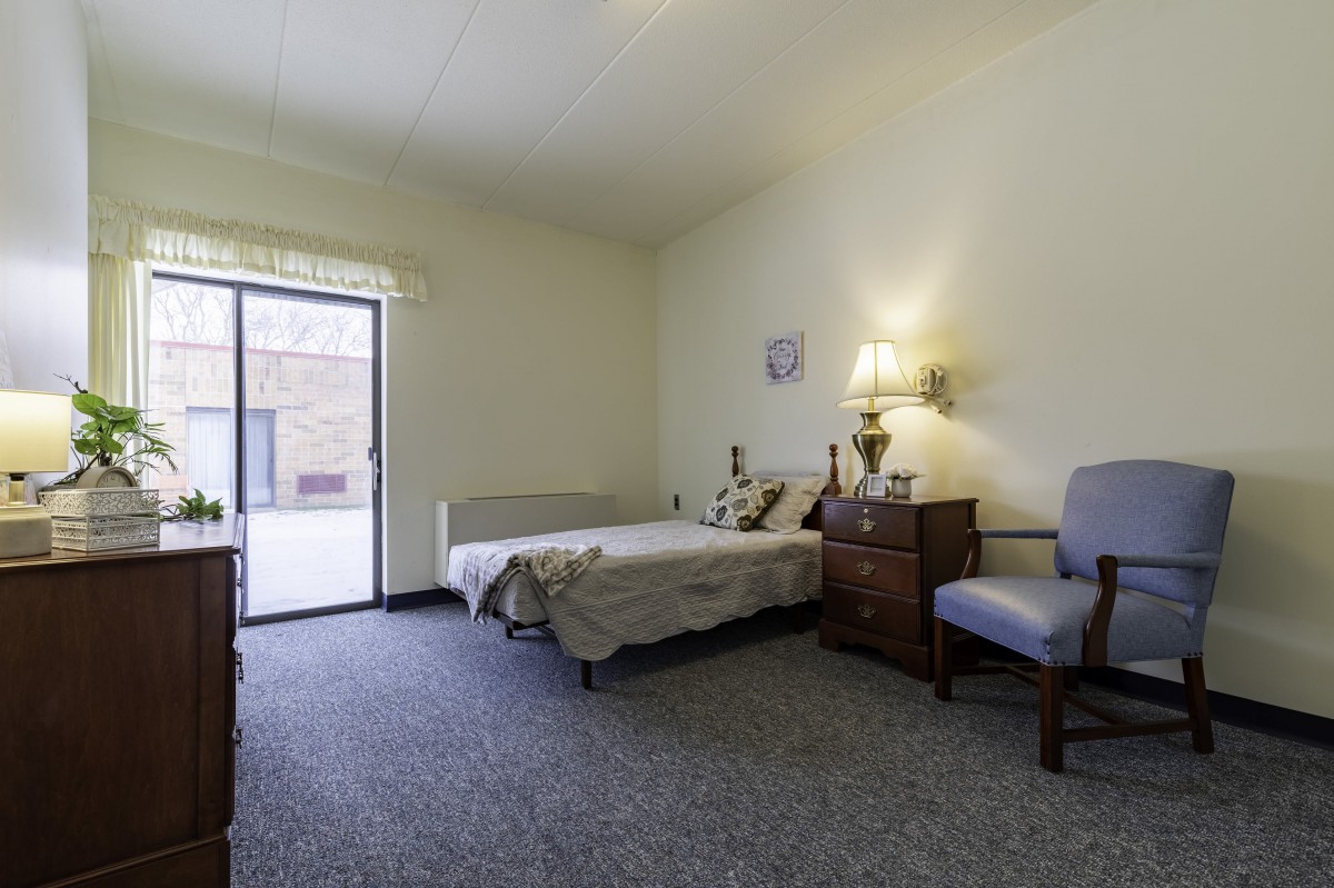 A minimalist bedroom with a bed, chair, and drawer under soft lighting.