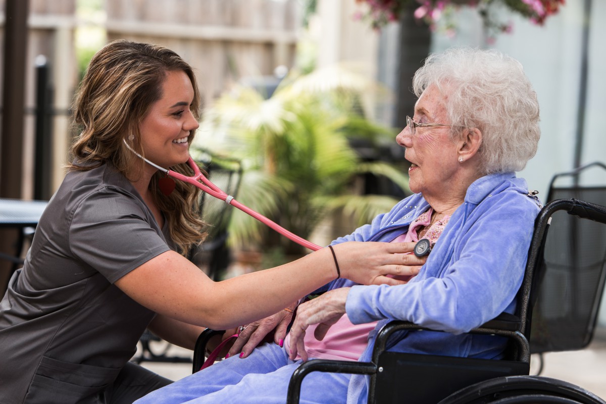 A nurse attentively checks the heart rate of an elderly patient seated in a wheelchair outdoors.