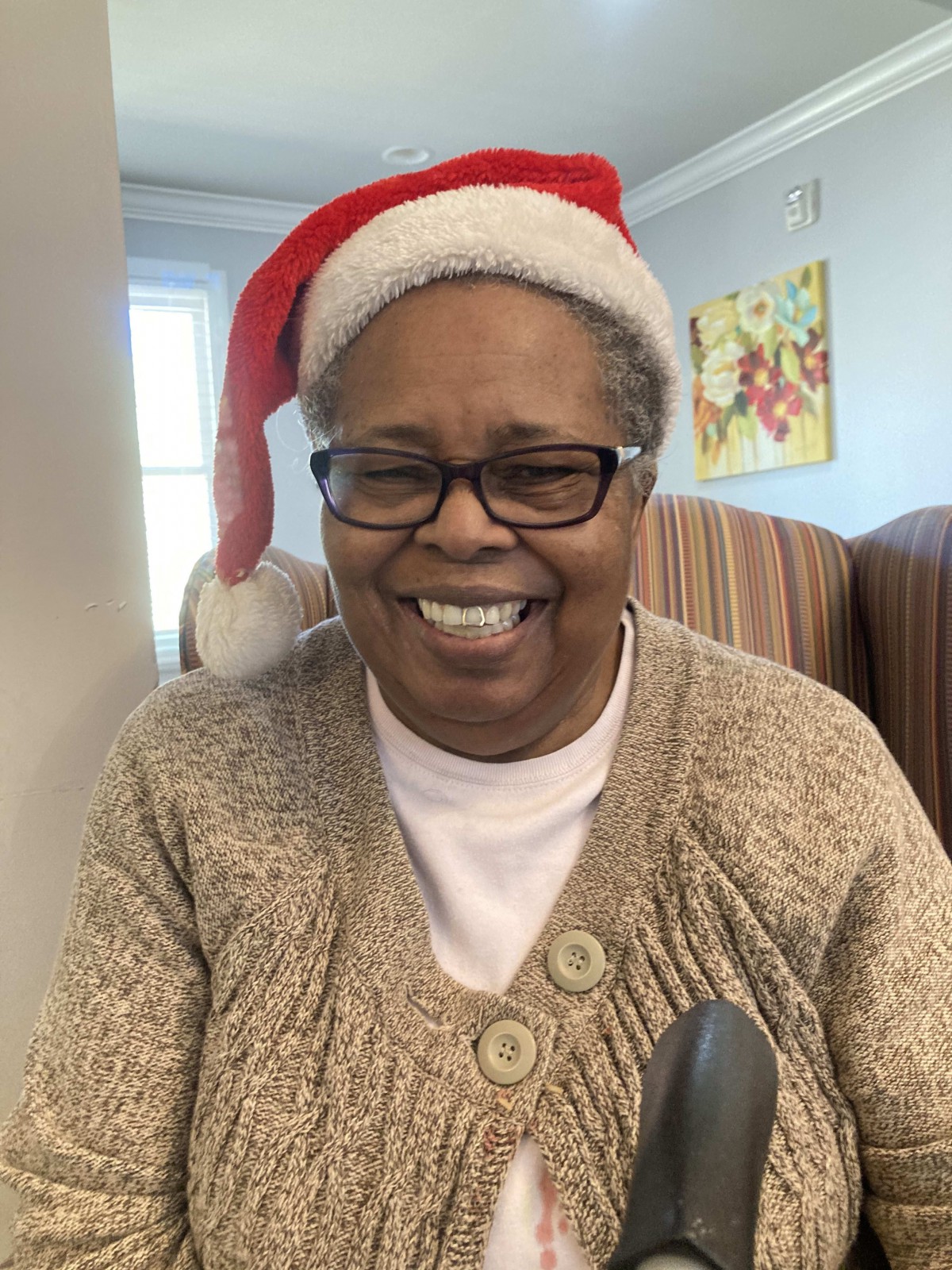 An elderly woman smiling while wearing a Santa hat and glasses indoors.