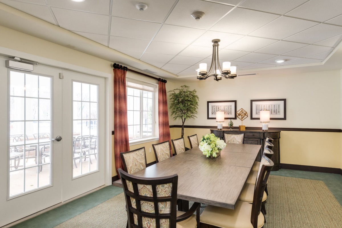 A spacious conference or dining room with a long table and several chairs, illuminated by a chandelier and decorated with artwork and plants.