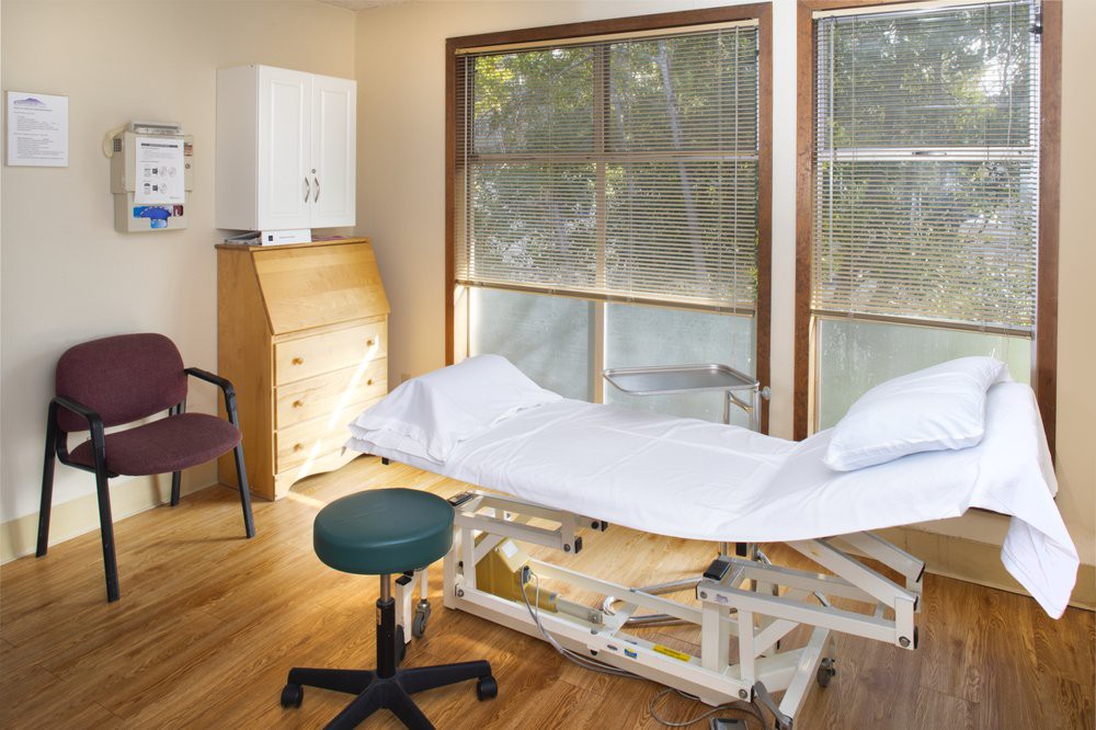 A clean and bright medical examination room with a table, chair, and windows.