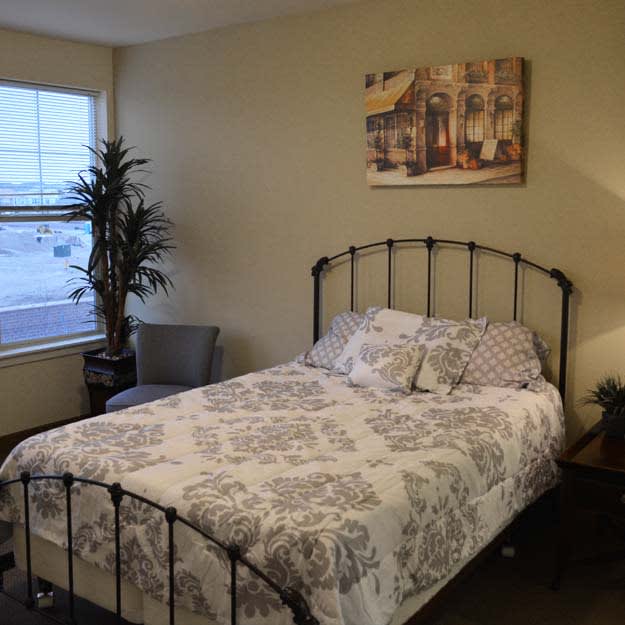 A cozy bedroom featuring a bed with floral bedding, artwork on the wall, a chair by the window, and a potted plant.