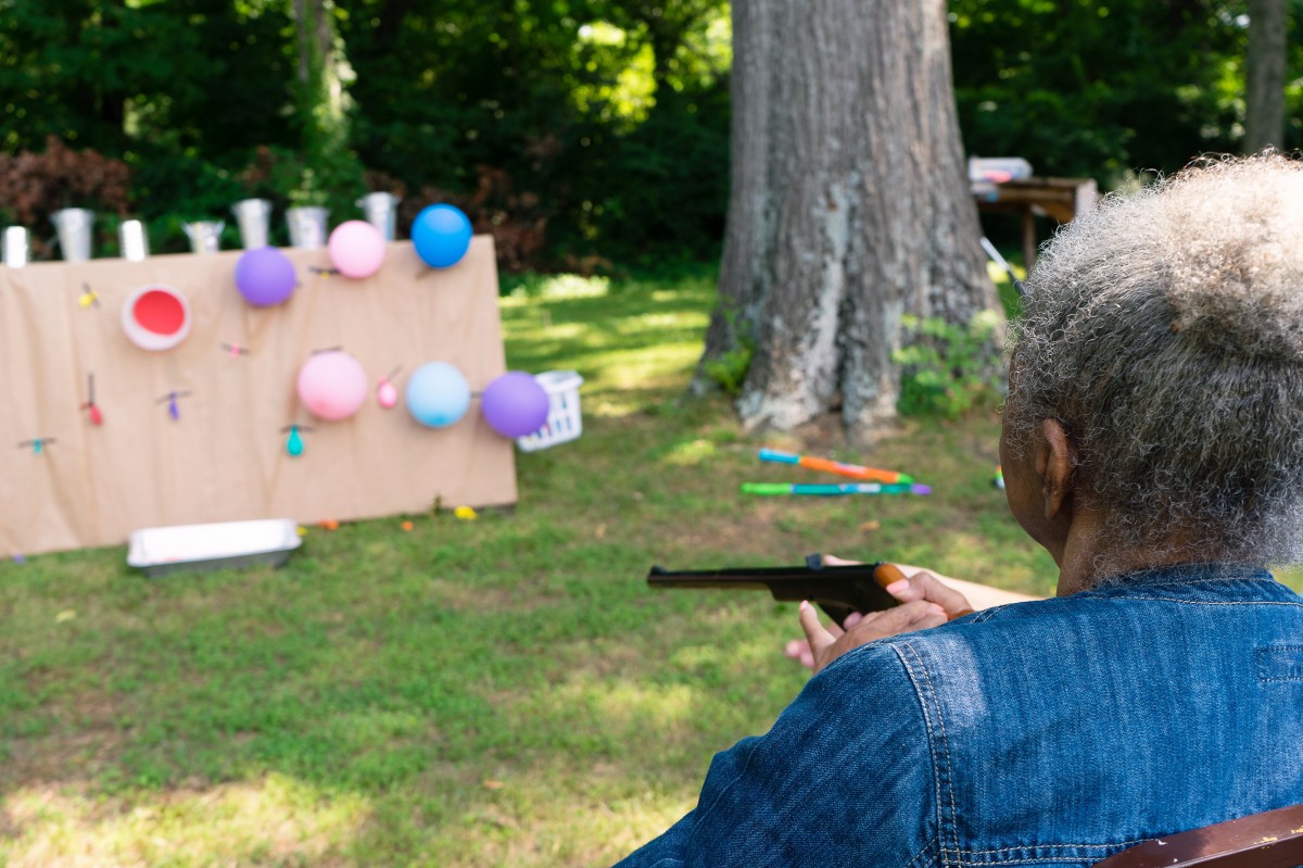 A person aims a pistol at a backyard target setup with colorful balloons.