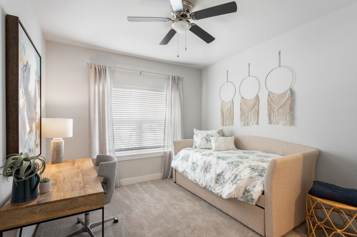 A cozy bedroom featuring a daybed, desk, and ceiling fan with soft lighting and subtle wall decor.