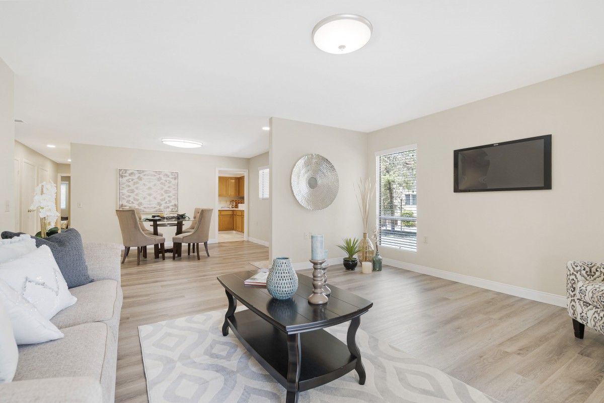 A modern and spacious living room with contemporary furniture and bright lighting, featuring wood flooring and a dining area in the background.