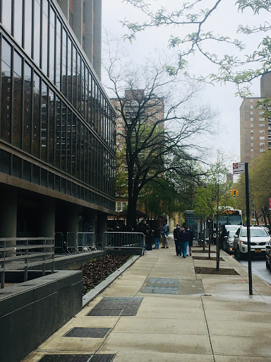 A bustling urban scene featuring pedestrians on a sidewalk next to modern buildings and greenery.
