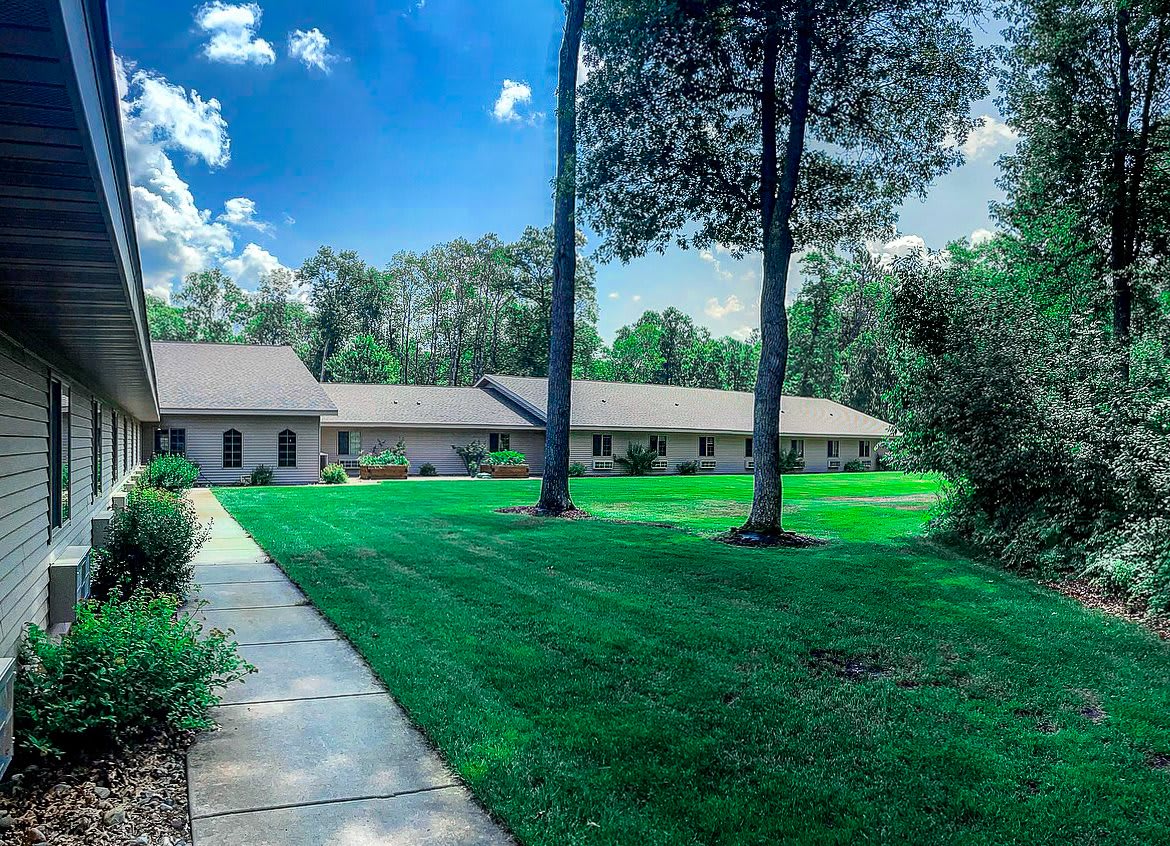 A scenic view of a house with a well-maintained lawn and tall trees against a bright blue sky.