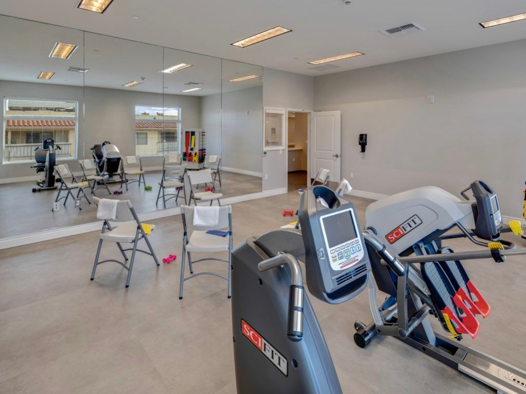 A fitness room with exercise machines and chairs, featuring large mirrors on the wall.