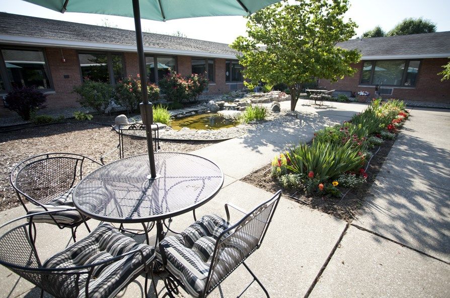 A serene patio area with a metal table, chairs, and an umbrella, surrounded by a lush garden.
