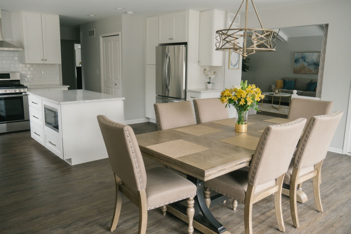 A modern dining area with a wooden table and cushioned chairs, featuring a stylish chandelier and a vase of yellow flowers.