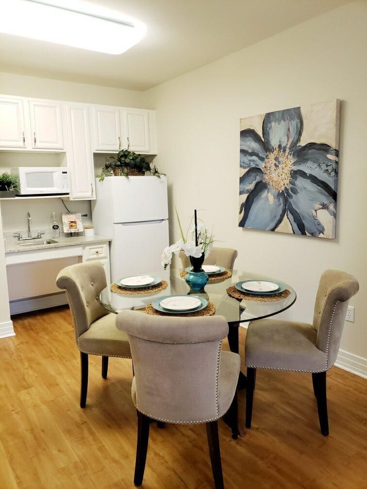 A cozy dining area with a round table set for four, adjacent to a compact kitchen featuring white cabinetry and appliances. A large flower painting adds a touch of elegance to the space.