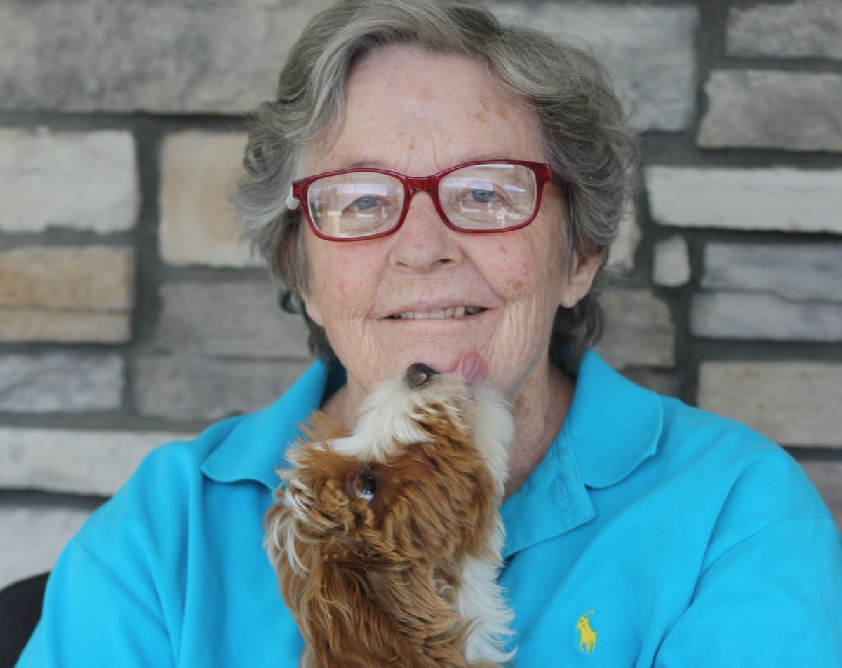 A person wearing glasses smiles while holding a small dog indoors.