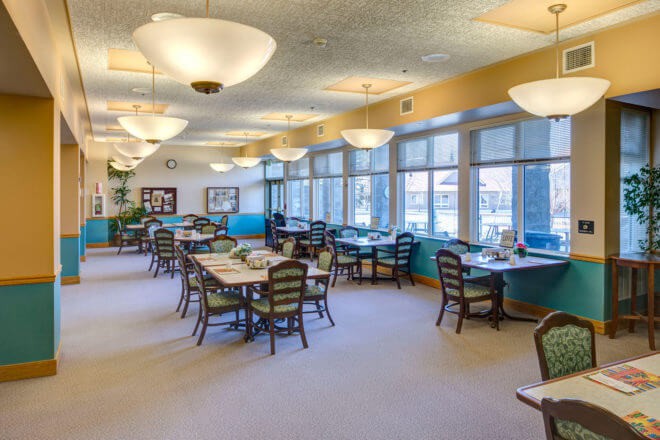 A spacious dining room with several tables and chairs arranged neatly, featuring large windows and elegant lighting fixtures.