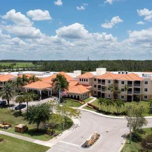An aerial view of a large building complex surrounded by palm trees and lush landscape under a partly cloudy sky.