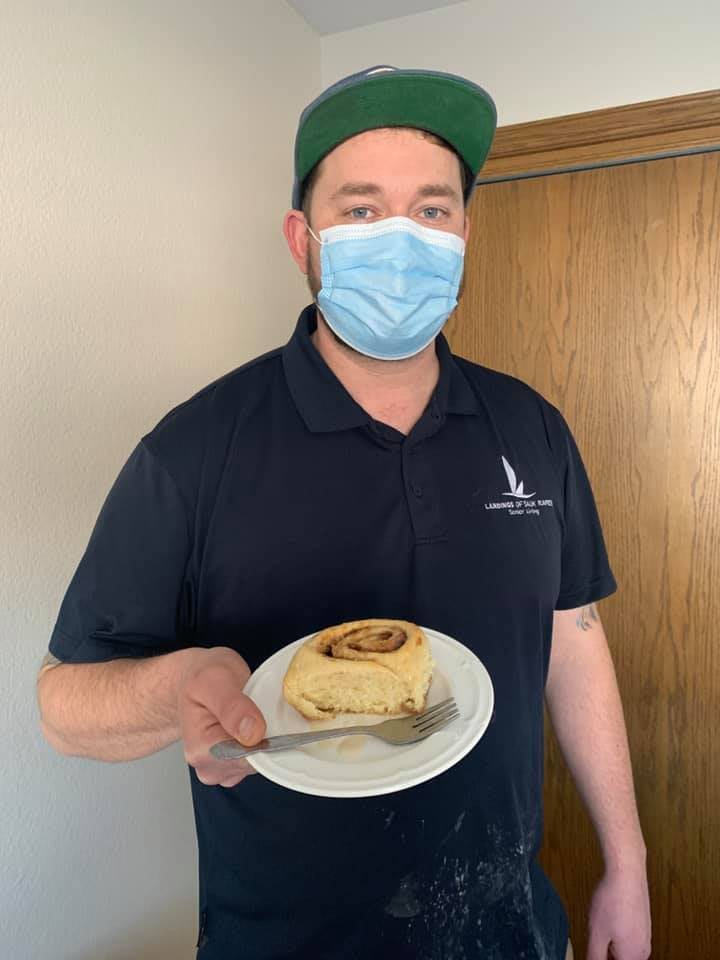 A person wearing a mask and hat holding a plate with a cinnamon roll.