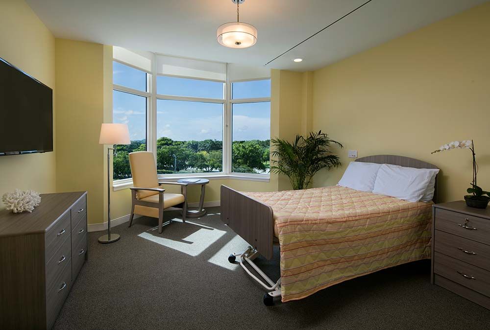 A well-lit hospital room with a patient bed and large windows providing natural light.