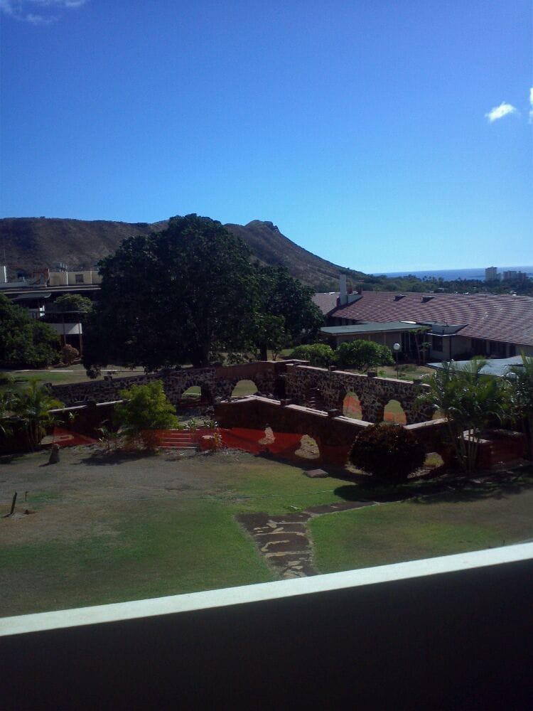 A scenic outdoor view featuring a grassy area with trees, buildings, and a hill under a clear blue sky.