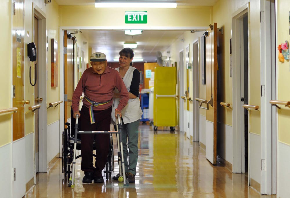 An elderly person using a walker is assisted by a caregiver in a nursing home hallway.