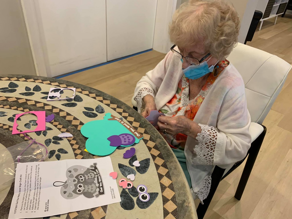 An elderly woman wearing a mask is engaged in a crafting activity, working with colorful paper cutouts at a table.