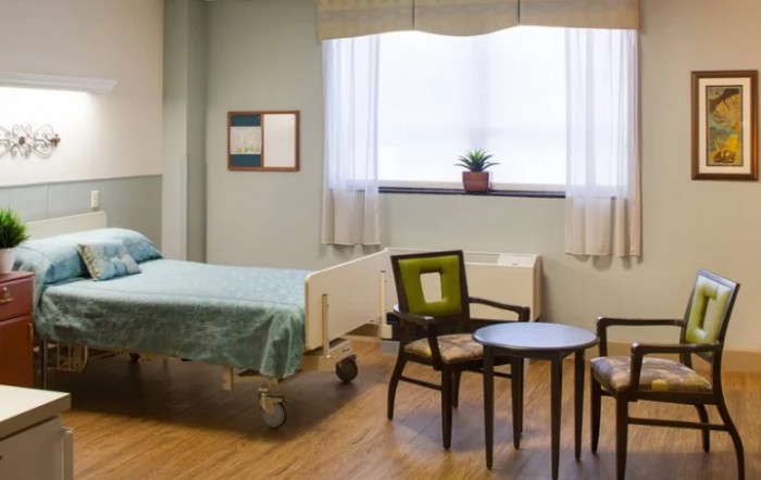 A clean and tidy hospital room with a bed, chairs, and a small table by the window.