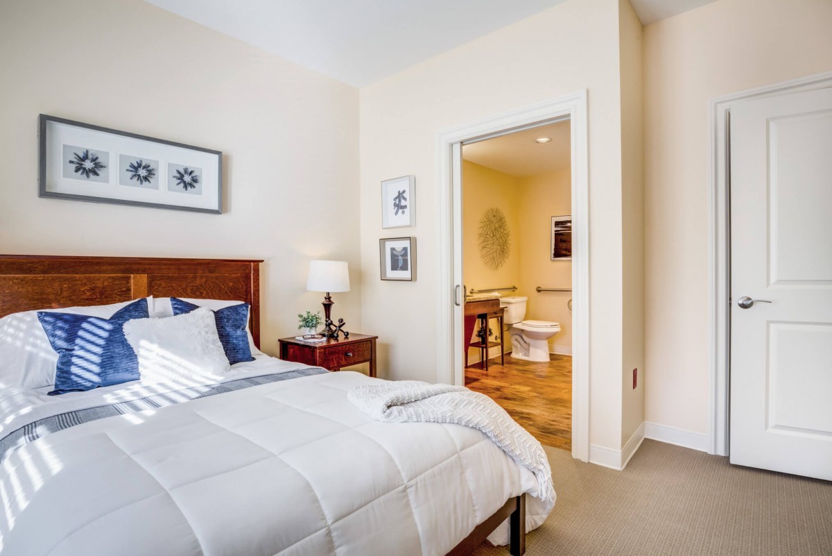 A cozy bedroom with a neatly made bed, bedside table, and an open doorway leading to a bathroom.