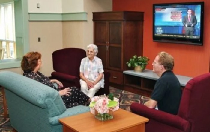 Three people are sitting in a living room, engaged in conversation with a television on the wall.