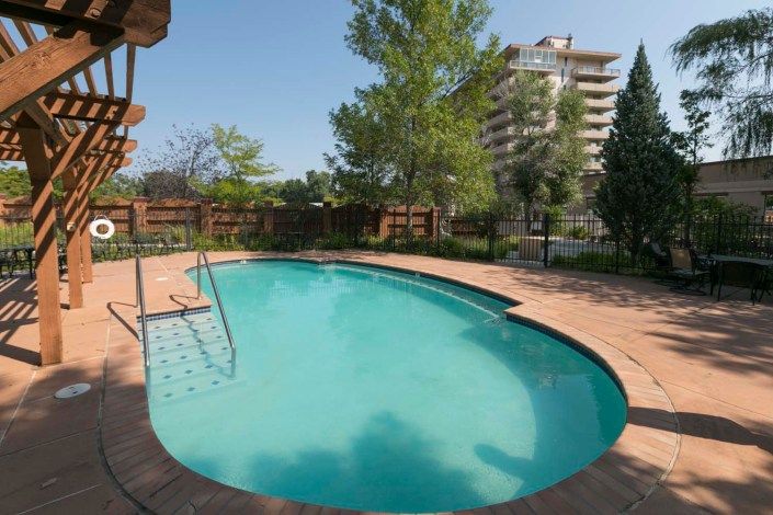 A tranquil outdoor swimming pool surrounded by trees and fencing, with a building visible in the background.