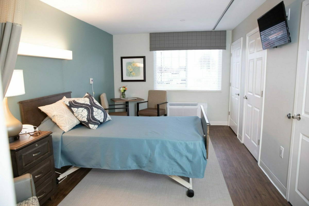 A modern and minimalist bedroom with a hospital bed, featuring soft blue bedding, a chair, and a large window.