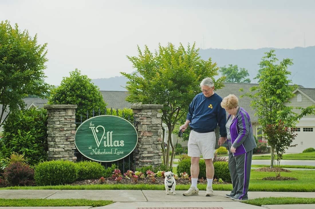 An elderly couple walking their dog near the entrance of a retirement community.