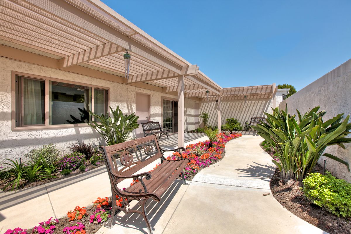 A well-maintained garden area with benches and a pergola, surrounded by vibrant flowers and plants along a paved pathway.