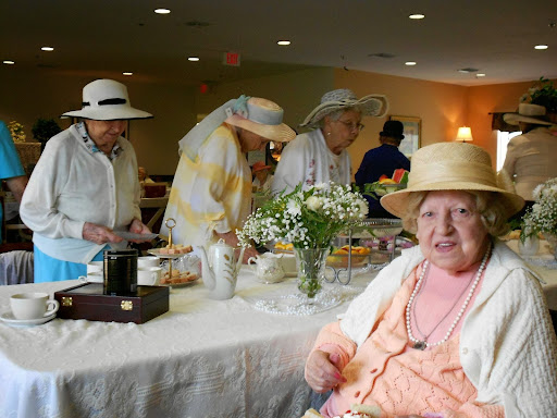 A joyful gathering of elderly women enjoying a tea party, dressed in hats and surrounded by flowers.