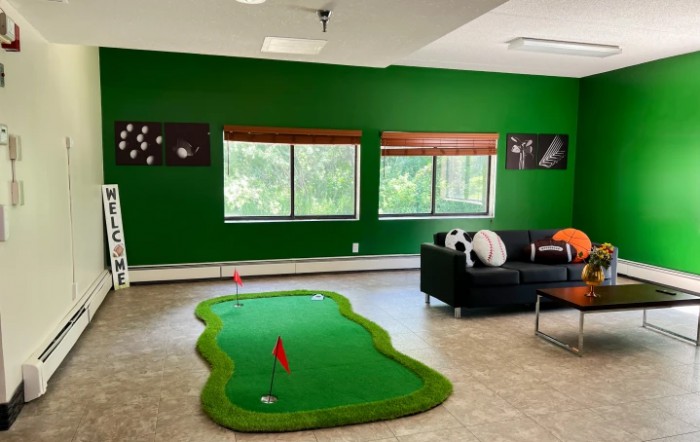 A bright recreation room with a mini golf setup, featuring green walls and large windows.