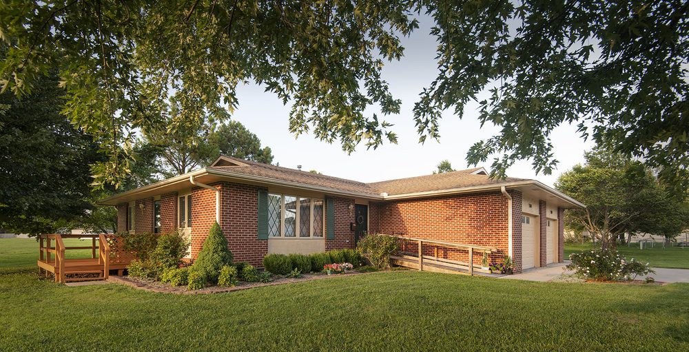 A suburban brick house with a front ramp and lush green lawn surrounded by trees.