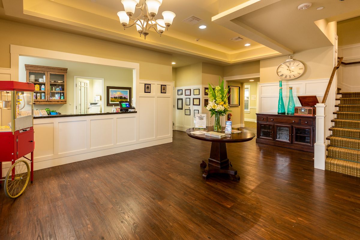 A warmly lit lobby with wooden flooring, featuring a reception desk and decorative elements like a chandelier and flower arrangements.