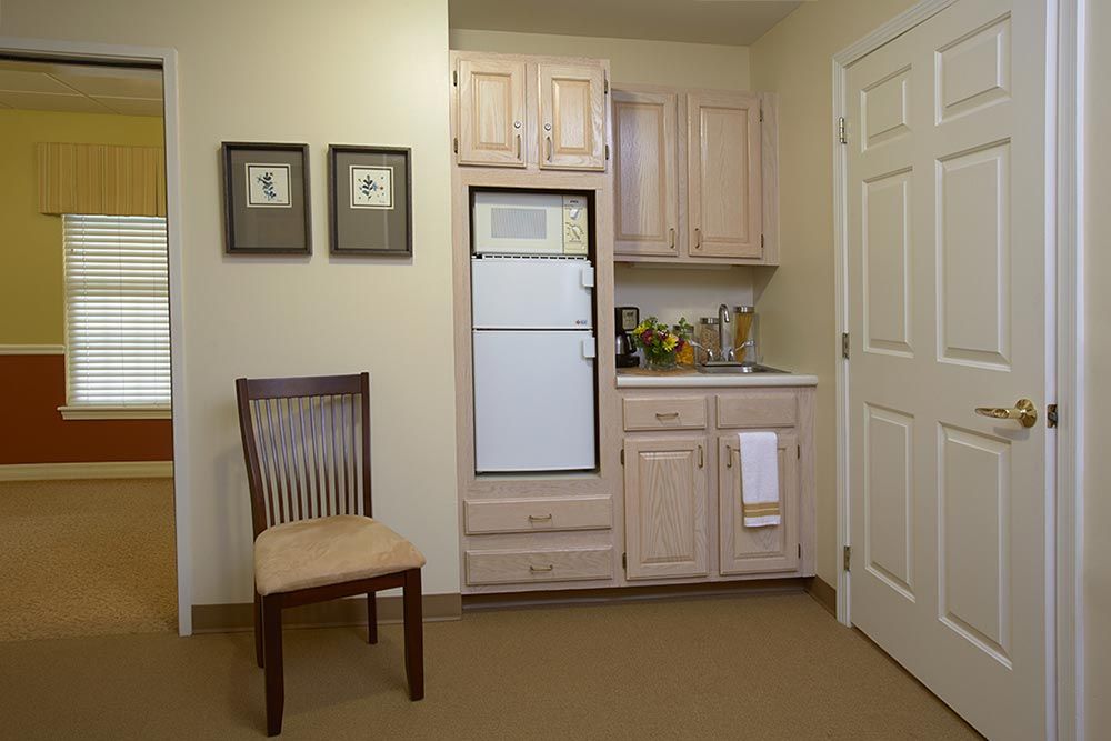 A compact kitchenette with light wood cabinets, a small fridge, and a chair in the corner.