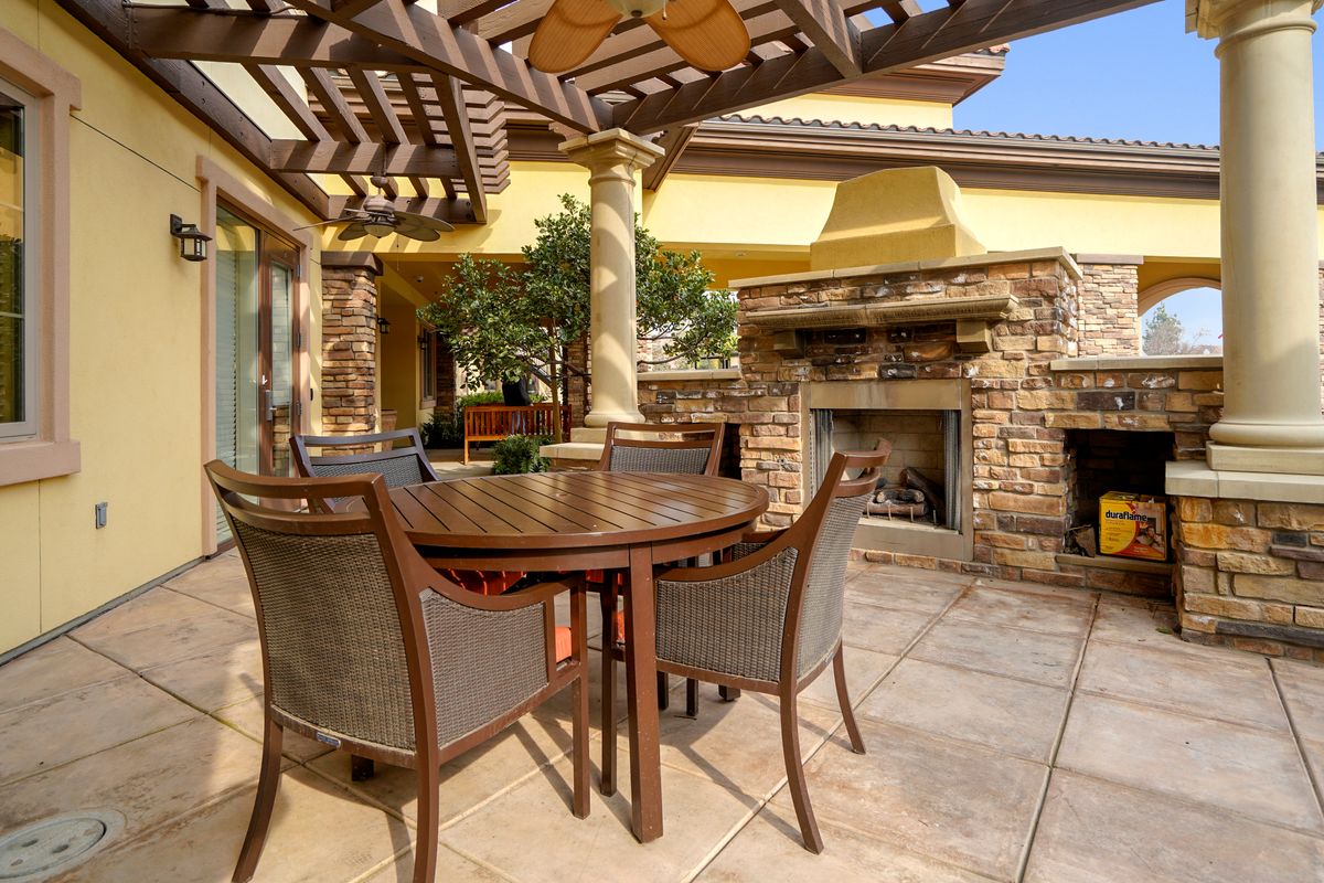 A sunny patio featuring an outdoor dining set under a pergola, with a stone fireplace in the background.