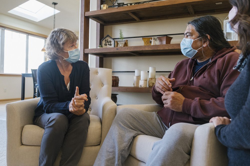 Two people wearing masks have a conversation in a living room setting.